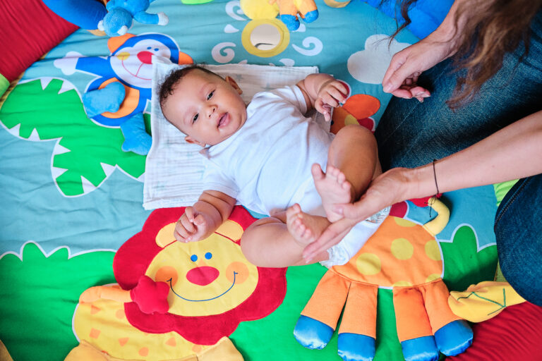 Top view of a baby lying on colorful play mat on the floor while playing with his mother at home.