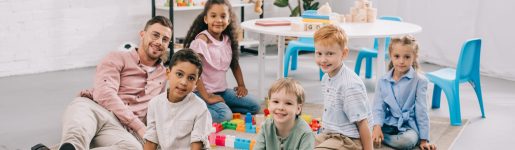 smiling teacher and multicultural preschoolers sitting on floor with colorful bricks in classroom
